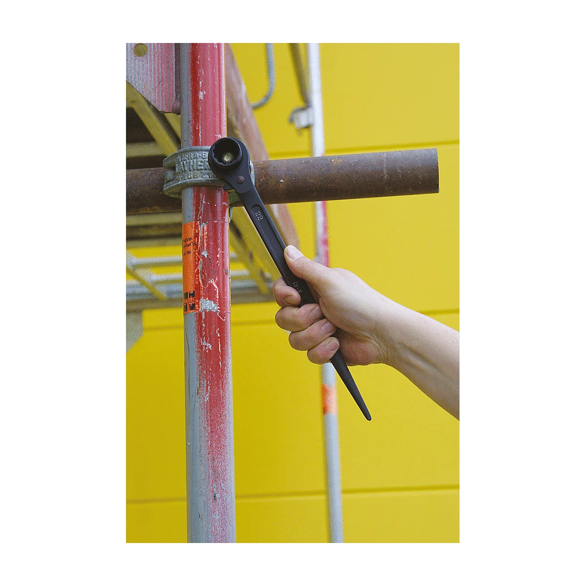 Eine Hand hält einen schwarzen Schraubenschlüssel an einer roten Gerüststange.